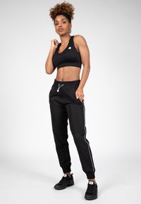 Zwart atletisch crop top met een logo op de borst, gecombineerd met zwarte joggers met witte zijstrepen en elastische manchetten, gedragen met zwarte sneakers.