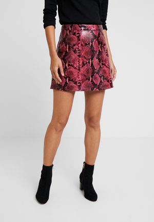 Femme portant une mini-jupe imprimé serpent rouge et noir, un top noir à manches longues et des bottines noires à talons blocs sur fond blanc.