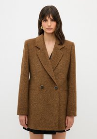 Ruskea teksturoitu blazer, jossa on kaksirivinen etuosa, terävät kaulukset ja kaksi nappia. Siinä on pitkät hihat ja sivutaskut.