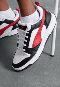 Puma-sneakers med en övre del i svart läder och vita samt röda accentfärger, en vit sula och perforerad tå för ventilation. Vita snören.