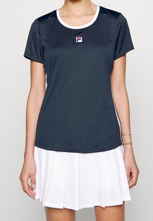 Kobieta ubrana w granatowy sportowy T-shirt z krótkim rękawem z małym logo oraz białą plisowaną spódnicę na jednolitym tle.