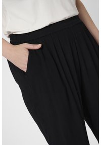 Personne portant un pantalon noir texturé avec une ceinture élastique, main reposant dans la poche avant, associé à un haut blanc.