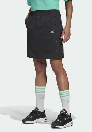 Person iført en sort Adidas nederdel med sidelås-knapper, hvide Adidas sokker med grønne striber, sorte Adidas sneakers og en grøn top.