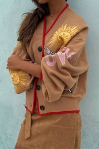 Beige cardigan med rød kant og stort solbroderi; har sorte knapper og oversized ærmer. Bæres over en beige suedepetit skirt.