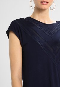 Kvinna i en marinblå ärmlös topp med spets- och meshdetaljer i chevronmönster, tillsammans med stora hoopörhängen, sedd från axeln till munnen.