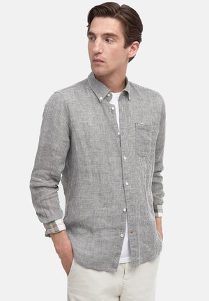 Mann mit Händen in den Taschen, trägt ein grau kariertes Hemd über einem weißen T-Shirt und schaut nach links gegen einen schlichten Hintergrund.