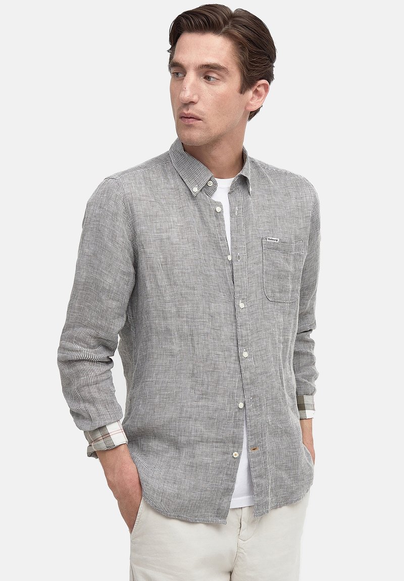 Mann mit Händen in den Taschen, trägt ein grau kariertes Hemd über einem weißen T-Shirt und schaut nach links gegen einen schlichten Hintergrund.