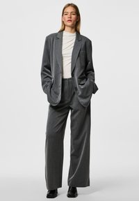 Blazer grigio doppiopetto con strisce verticali, abbinato a pantaloni a gamba larga coordinati. Tessuto morbido, vestibilità oversize e tasche frontali. Stivali neri.