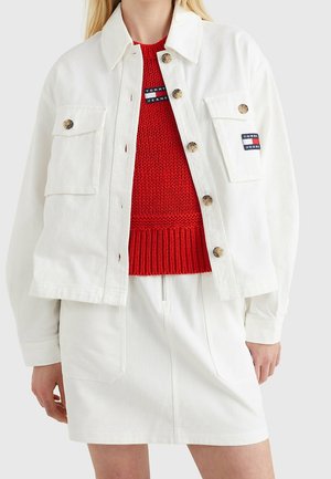 Persona vistiendo una chaqueta blanca con botones y bolsillos con solapa, falda blanca con cremallera frontal y suéter rojo de punto con logotipos de "Tommy Jeans".