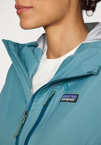 Jachetă teal din material ușor, cu fermoar frontal, glugă ajustabilă și patch cu logo-ul Patagonia pe piept.