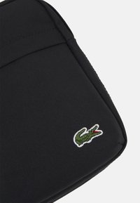Funda negra para portátil con tejido texturizado, que presenta un logo de cocodrilo bordado en verde en la esquina inferior derecha.