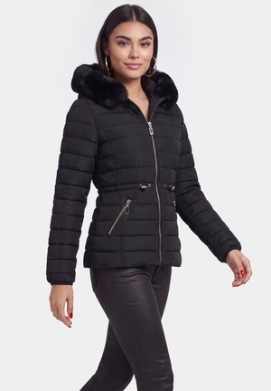Donna che indossa una giacca nera trapuntata con cappuccio bordato di pelliccia, tasche con zip e pantaloni neri in pelle, cammina su uno sfondo semplice.
