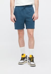 Shorts en coton bleu avec une ceinture élastique et un cordon de serrage. Portés avec des baskets montantes grises et des chaussettes blanches. Ourlets simples avec bande.