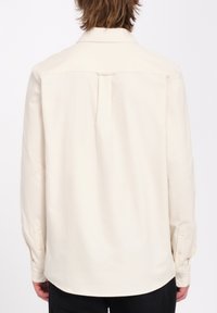 Chemise à manches longues beige clair avec un col classique, un pli simple au dos et des poignets à bouton. Tissu lisse avec un léger éclat.