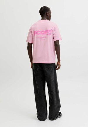 Uomo in piedi con la schiena rivolta, che indossa una t-shirt rosa con grafica e testo e jeans neri larghi su uno sfondo bianco semplice.