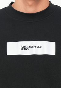 Čierna mikina so žebrovaným golierom a bielou obdĺžnikovou logom s nápisom "KARL LAGERFELD JEANS" v čiernom písmene.