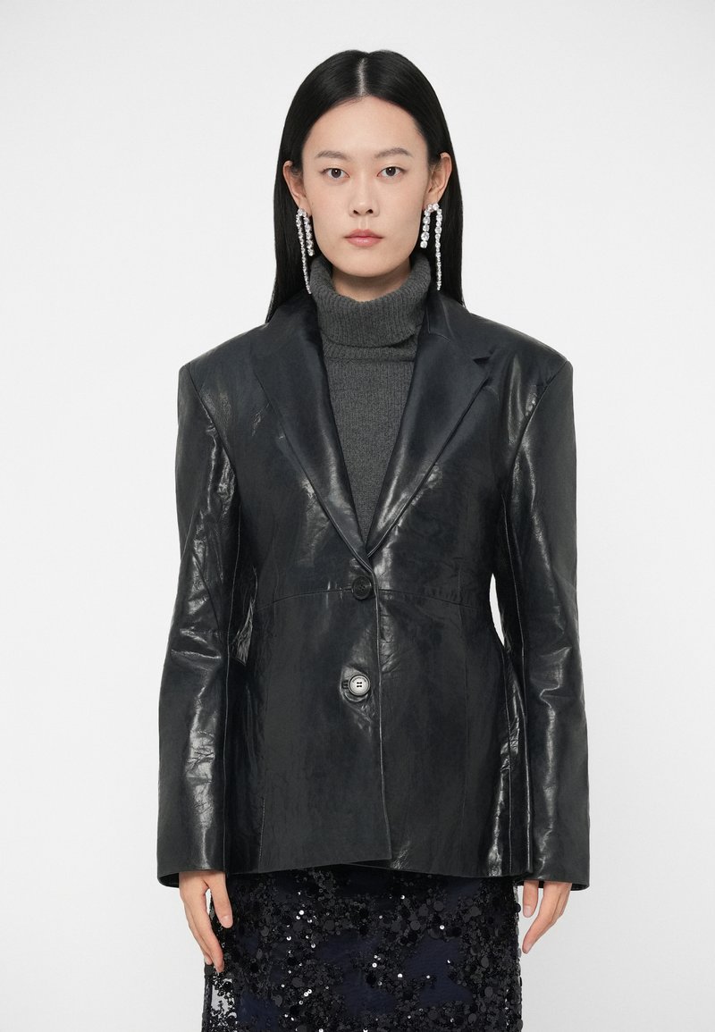 Blazer de couro preto com um design estruturado, apresentando um fecho de um único botão, lapelas pontiagudas e textura suave. Usado sobre um suéter de gola alta cinza.