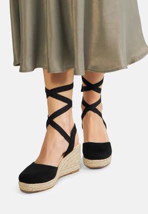 Czarne zamszowe buty na koturnie z plecionymi podeszwami z juty i krzyżującymi się materiałowymi paskami wokół kostek, zestawione z zwiewną oliwkowo-zieloną spódnicą.