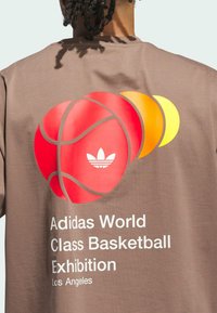 Hnedé bavlnené tričko s veľkým červeným grafikom basketbalu, oranžovými a žltými akcentmi a bielym textom "Adidas World Class Basketball Exhibition."