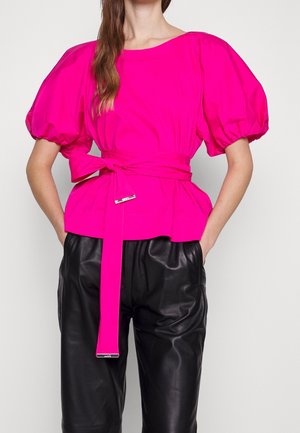 Frau trägt eine leuchtend rosa Bluse mit Puffärmeln und einem langen, gebundenen Gürtel, kombiniert mit lockeren schwarzen Lederhosen, die Hände in den Taschen.