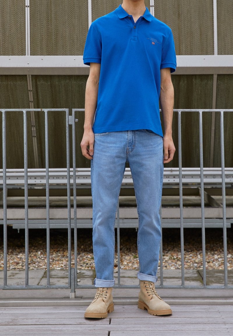 Homme portant un polo bleu, un jean clair retroussé et des bottines beige à lacets, debout devant une rambarde métallique.