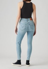 Frau in hellblauen hochtaillierten Skinny-Jeans, schwarzem Tanktop und schwarzen High-Top-Sneakern, die vor einem schlichten Hintergrund steht.