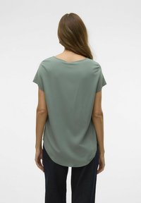 Camiseta de manga corta en verde sutil, hecha de una tela suave con un dobladillo trasero redondeado y un escote abierto, combinada con pantalones de mezclilla oscuros.