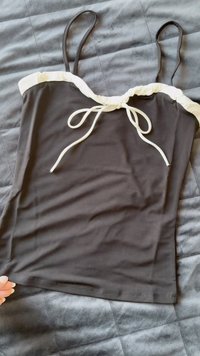 Black tank top with thin straps, a white ruffled neckline, and a front tie detail. Smooth fabric and fitted shape.