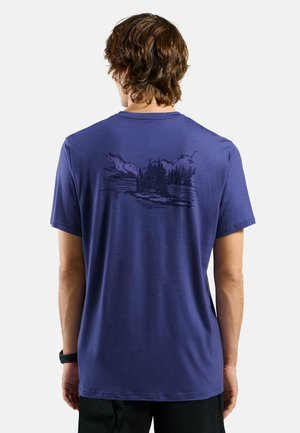 Mann trägt blaues T-Shirt mit schwarzer Berg- und Waldlandschaftsgrafik auf dem oberen Rücken, steht vor schlichtem weißem Hintergrund.