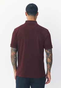 Polo color bordeaux con motivo a rilievo, maniche corte e colletto. Presenta polsini a coste e orlo dritto.