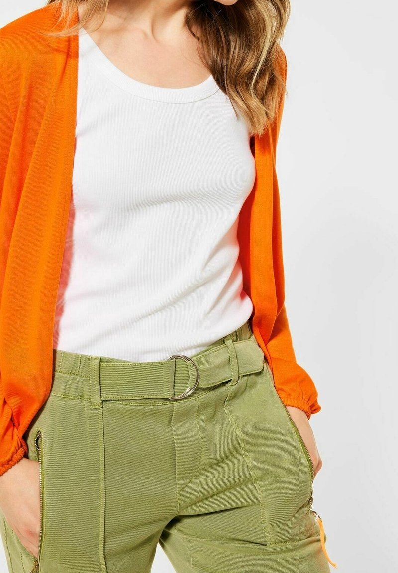 Světlý oranžový lehký cardigan přes bílý žebrovaný top, doplněný olivově zelenými cargo kalhotami se stříbrným řetízkem a bočními zipy.