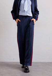Persona vistiendo pantalones de pierna ancha de color azul marino con rayas rojas a los lados, una camisa de color azul claro, una chaqueta azul marino y mocasines negros, de pie con las manos en los bolsillos.