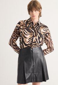 Camicia con bottoni in stampa animale con tonalità di marrone e nero, abbinata a una gonna in pelle nera aderente con cerniera frontale e pieghe.
