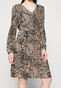 Femme portant une robe beige mi-longue à manches longues avec des motifs animaux noirs mélangés, debout devant un fond uni.