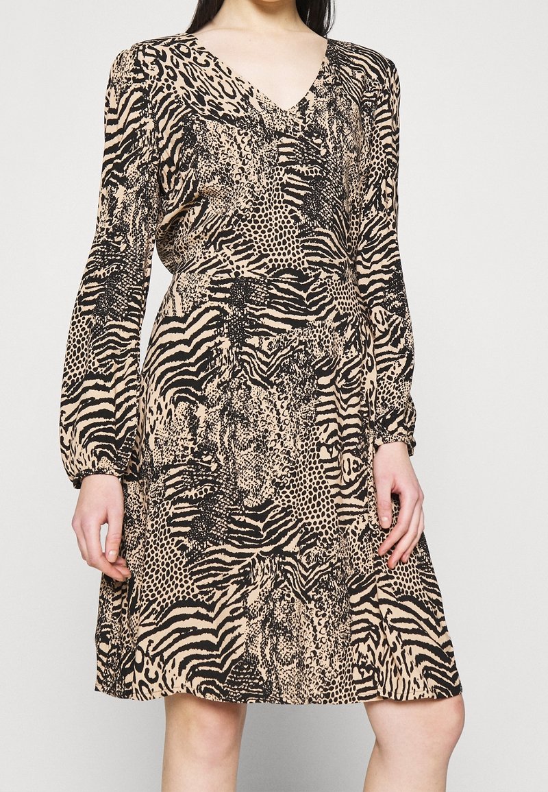 Femme portant une robe beige mi-longue à manches longues avec des motifs animaux noirs mélangés, debout devant un fond uni.