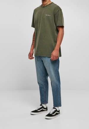 Olivgrünes Baumwoll-T-Shirt mit Rundhalsausschnitt und kurzen Ärmeln, kombiniert mit hellblauen Straight-Leg-Jeans und schwarzen und weißen Canvas-Sneakers.