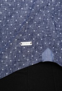 Blauwe stofshirt met een witte bloemenprint, een label met de tekst "EDC BRAND", en een gestreepte detail aan de binnenrand.