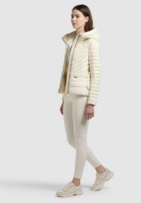 Femme portant une veste matelassée crème à capuche, un legging assorti et des baskets blanches, debout avec une jambe légèrement pliée sur un fond uni.