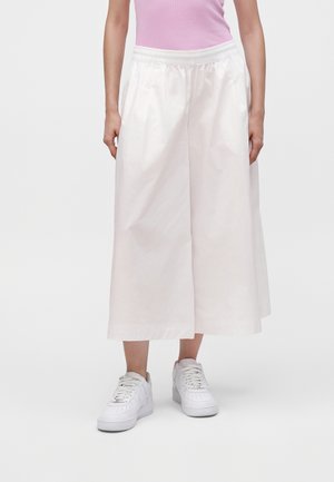 Femme portant un pantalon blanc large court, des baskets blanches et un haut rose sans manches, debout devant un fond uni.