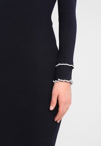 Niebieska sukienka z prążkowanej tkaniny, o dopasowanym kroju, z białymi falbankami przy mankietach, wykonana z miękkiego i teksturowanego materiału.