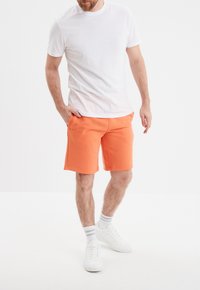 Pantalones cortos de algodón naranja con bolsillos, que presentan un corte recto y un dobladillo. Combinados con una camiseta básica blanca de cuello redondo y zapatillas blancas.