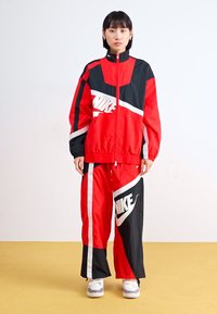 Rdeča in črna Nike trenirka, ki vsebuje jakno z zadrgo in široke hlače. Drzni beli logotipi, geometrijski barvni bloki in lahka tkanina.