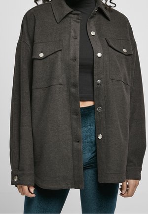 Veste boutonnée gris foncé à poches poitrine, portée ouverte sur un top court noir et un pantalon en velours turquoise sur une personne aux cheveux bouclés.