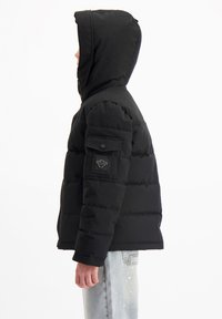 Zwarte pufferjack met capuchon, een ritssluiting aan de voorkant en een zijzak met klep. Textuur met een gewatteerd ontwerp en een strakke pasvorm.