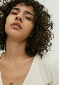 Model met krullend donker haar in een lichtbeige ribgebreide top met v-hals. Opvallende details zijn de zichtbare huidtextuur en een kleine tatoeage op de nek.