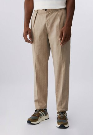 Mężczyzna ubrany w beżowe spodnie w prążki oraz brązowe, zielone i czarne sportowe buty, stojący na prostym tle.