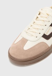 La sneaker presenta una toma in pelle color crema con punta in suede marrone, dettagli beige, lacci color crema e suola in gomma testurizzata con scanalature.