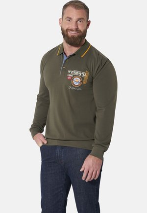 Man with beard wearing green long-sleeve polo shirt with yellow details and patches, and dark blue jeans, smiling with one hand in pocket.