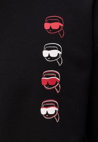 Tessuto nero con quattro volti stilizzati ricamati che indossano vari occhiali da sole. I colori includono rosso e bianco come accenti nei disegni.