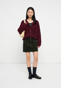 Jovem mulher vestindo um suéter bordô com botões, saia verde-oliva com bolsos, meias escuras, sapatos pretos e carregando uma bolsa de ombro bege, de pé.